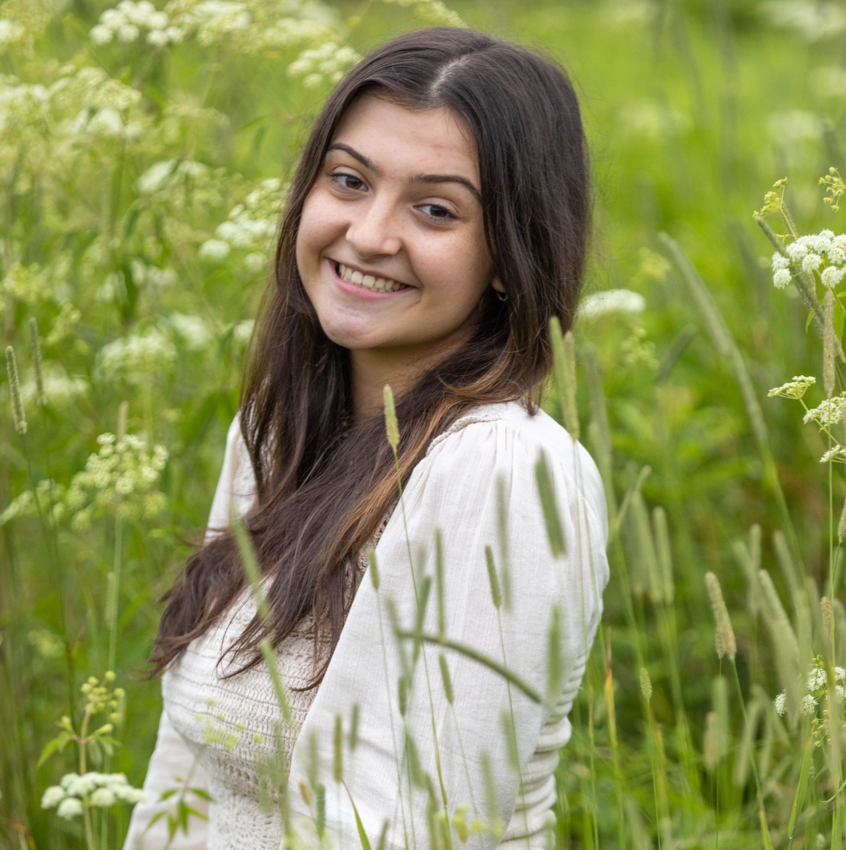 A portrait of the creator of the website. A white woman with long dark hair. She is sitting in a green field and wearing a white shirt.